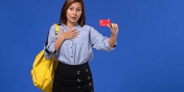 front-view-young-female-blue-shirt-wearing-yellow-backpack-holding-red-plastic-card-blue-wall