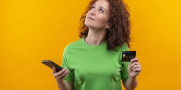 young woman with short curly hair in green t-shirt holding two smartphones looking up with pensive expression on face trying to make choice standing over orange background